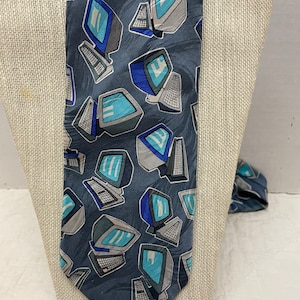 Puede incluir: Una corbata azul oscuro con un patrón repetido de monitores de computadora retro en tonos de gris, azul y verde azulado. La corbata se exhibe en un maniquí de color neutro. El diseño incluye teclados y las pantallas de los monitores.