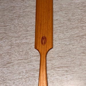 May include: A wooden spatula with a long, rectangular blade and a rounded handle. The spatula is made of light brown wood and features a circular logo near the blade. A hole is drilled at the end of the handle.