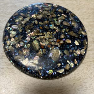 Puede incluir: Posavasos redondo negro con conchas iridiscentes y purpurina dorada incrustadas. El posavasos tiene el texto "VIRGINIA BEACH VIRGINIA" en letras doradas.