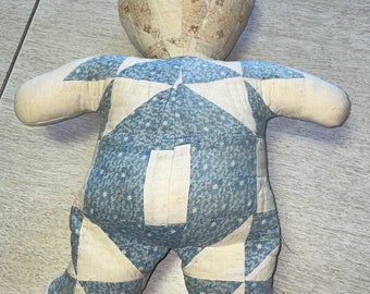 May include: A handmade teddy bear made from quilted fabric. The bear's body is a patchwork of blue and white diamond patterns, while the head and limbs feature a floral print. The ears have red and blue stripes.