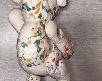 May include: A handmade teddy bear with a patchwork design. The bear's body is primarily white, with patches of fabric featuring floral and geometric patterns in various colors, including green, yellow, and red. The bear has black button eyes.
