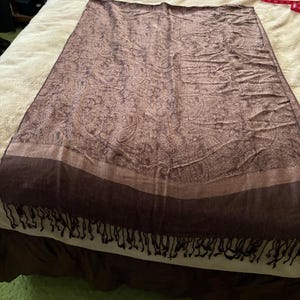 May include: A brown paisley patterned scarf with a dark brown border and fringe. The scarf is rectangular and appears to be made of a soft material. The paisley pattern is visible throughout the scarf.