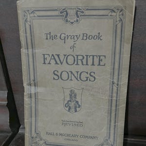 May include: The Gray Book of Favorite Songs, a vintage songbook with a gray cover and blue lettering. The cover features decorative borders and the text "REVISED" and "HALL & McCREARY COMPANY CHICAGO". The book is slightly worn, suggesting its age.
