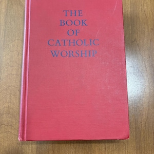 May include: A red hardcover book titled "THE BOOK OF CATHOLIC WORSHIP" in blue lettering. The book is rectangular and appears to be in good condition, resting on a wooden surface.