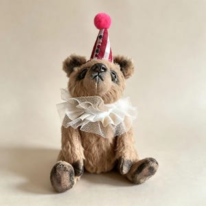 May include: A light brown teddy bear with dark-tipped fur on its paws and nose. The bear wears a white ruffled tulle collar around its neck and a pointed party hat featuring a dark red and white pattern topped with a small bright pink pom-pom.