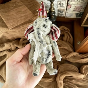 May include: A small, handcrafted elephant figurine made of light gray textured fabric with visible dark stitching. It wears a red and white striped cone hat topped with a red gem and gold beads, and features matching striped ears and a white fabric collar.