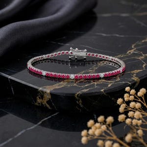 Puede incluir: Una pulsera de tenis en tono plateado con una hilera continua de piedras redondas de color rojo vibrante intercaladas con piedras blancas transparentes. La pieza incluye un cierre de caja metálico seguro, con un diseño refinado y elegante.