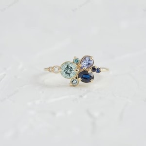 Könnte beinhalten: Ein goldener Ring mit einem Cluster aus blauen und grünen Edelsteinen. Der Ring hat einen zentralen, runden, hellblauen Stein, umgeben von kleineren Steinen in verschiedenen Formen und Farbtönen. Das Band ist dünn und goldfarben.