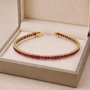 Könnte beinhalten: Ein goldenes Tennisarmband mit einer durchgehenden Reihe quadratisch geschliffener, leuchtend pink-roter Edelsteine, die in einen polierten Goldmetallrahmen gefasst sind. Das Armband verfügt über einen sicheren Metallverschluss am Ende.
