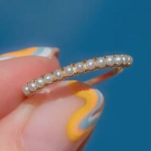 Op de afbeelding: Een delicate gouden ring versierd met een rij kleine, ronde parels. De ring wordt tegen een blauwe achtergrond gehouden, wat het elegante ontwerp benadrukt. De parels zijn gelijkmatig verdeeld, wat een klassieke en tijdloze uitstraling creëert.