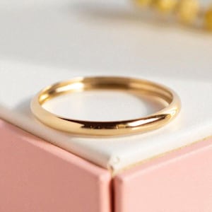 May include: A thin, polished gold band ring with a smooth, rounded surface. The ring features a continuous, circular design with a reflective metallic finish that catches the light, showcasing a simple, unadorned, and uniform gold construction.