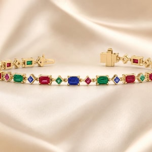 May include: A gold link bracelet featuring a repeating pattern of rectangular and square-cut gemstones in vibrant shades of red, green, and blue. The bracelet is constructed with a secure box clasp mechanism and polished metal settings.
