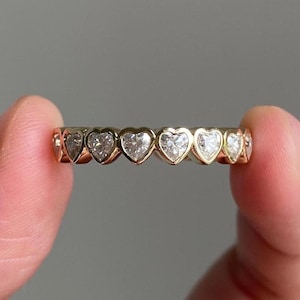 Könnte beinhalten: Ein goldener Ring mit einer durchgehenden Reihe von in Zargenfassung gefassten, herzförmigen, klaren Steinen. Der Ring besteht aus einer polierten Fassung aus gelbem Gold, die jeden einzelnen herzförmigen Stein in einem Muster um den Ring rahmt.
