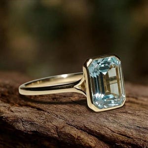 Könnte beinhalten: Ein Goldring mit einem großen, rechteckigen, smaragdgeschliffenen hellblauen Edelstein in einer glatten Zargenfassung. Das Band besteht aus poliertem Gold und der Stein weist klare Facetten auf, die das Licht reflektieren und einen blassblauen Farbton zeigen.