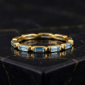 Könnte beinhalten: Ein goldener Ring mit einer durchgehenden Reihe hellblauer, im Baguetteschliff geschliffener Edelsteine, die horizontal angeordnet sind. Das Metallband hat eine polierte Oberfläche und die Steine sind rundherum aneinandergereiht.