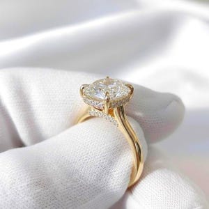 Op de afbeelding: Een gouden ring met een grote, gefacetteerde middensteen in kussenslijpvorm. De zetting bevat een halo van kleine, heldere steentjes rond de basis van de middensteen en extra kleine steentjes ingelegd langs de bovenkant van de ringband.