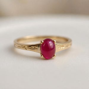 Könnte beinhalten: Ein goldener Ring mit einem glatten, ovalen, tiefrosa Cabochon-Edelstein in einer Vier-Krappen-Fassung. Der Goldring ist mit einem zarten, sich wiederholenden Blattmuster graviert, das sich auf beiden Seiten der zentralen Steinfassung erstreckt.