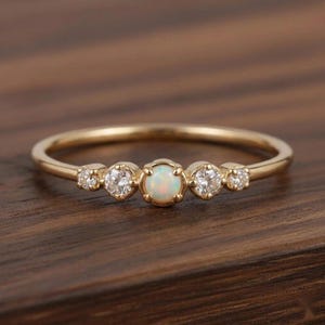 Könnte beinhalten: Ein goldener Ring mit einem zentralen, runden Opal-Cabochon, der auf jeder Seite von zwei kleinen, runden, klaren Diamanten flankiert wird. Der Ring ist dünn und poliert und zeigt ein filigranes Design mit insgesamt fünf horizontal gesetzten Steinen.