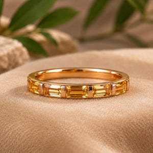 Könnte beinhalten: Ein goldener Ring mit einem Band aus rechteckigen gelben Edelsteinen. Der Ring liegt auf einer beigen Oberfläche mit einer verschwommenen grünen Pflanze im Hintergrund. Die Edelsteine sind gleichmäßig verteilt.