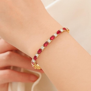 Könnte beinhalten: Ein goldfarbenes Armband mit einem abwechselnden Muster aus rechteckig geschliffenen roten Edelsteinen und rund geschliffenen klaren weißen Steinen. Das Schmuckstück verfügt über eine flexible Gliederkonstruktion, die sich um das Handgelenk legt und einen lebendigen Kontrast zwischen den tiefroten und den funkelnden klaren Steinen zeigt.