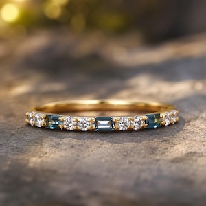 Pode incluir: Um anel de ouro com pedras preciosas claras e azuis alternadas. O anel apresenta pedras redondas e de corte baguette embutidas em uma faixa de ouro amarelo. O anel é exibido em uma superfície texturizada.