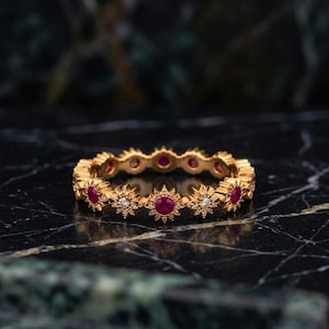 May include: A gold eternity band featuring alternating round red rubies and small clear diamonds set within star-shaped metal frames. The ring has a continuous circular design with a polished gold finish, showcasing a repeating pattern of gemstones.