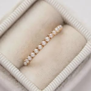 Puede incluir: Un delicado anillo de oro con una fila de pequeñas perlas blancas. El anillo está engastado en una banda de oro y se muestra dentro de un joyero suave y de color claro. Las perlas están espaciadas uniformemente.