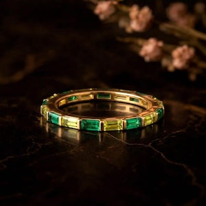 Könnte beinhalten: Ein goldener Ring mit abwechselnd grünen und gelben rechteckigen Edelsteinen. Der Ring liegt auf einer dunklen, reflektierenden Oberfläche, mit verschwommenen floralen Elementen im Hintergrund. Die Edelsteine sind Smaragd und Peridot.