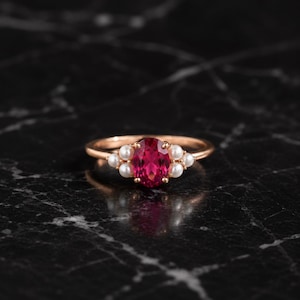 Könnte beinhalten: Ein Ring aus Roségold mit einem zentralen, oval geschliffenen, leuchtend pinken Edelstein, der auf jeder Seite von drei kleinen weißen Perlen flankiert wird. Die Ringschiene ist schmal und poliert und zeigt ein klassisches Design.