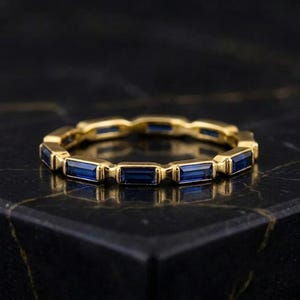 May include: A gold band ring featuring a continuous row of rectangular baguette-cut blue sapphires set in a channel setting. The metal has a polished finish, and the stones are arranged horizontally around the entire circumference of the ring.