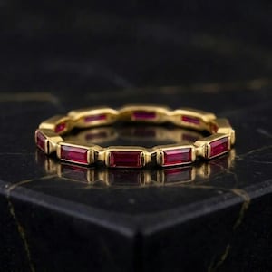 Könnte beinhalten: Ein Goldring mit einer durchgehenden Reihe rechteckiger, im Baguetteschliff geschliffener roter Rubine, die in einen polierten Metallrahmen gefasst sind. Der Ring zeigt ein sich wiederholendes Muster aus leuchtend roten Edelsteinen.