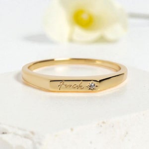 Könnte beinhalten: Ein goldfarbener Ring mit einem flachen Steg auf der Vorderseite. Der Steg ist mit dem kursiven Schriftzug "Fwck" graviert und mit einem kleinen, klaren, sternförmigen Stein rechts neben dem Schriftzug verziert.