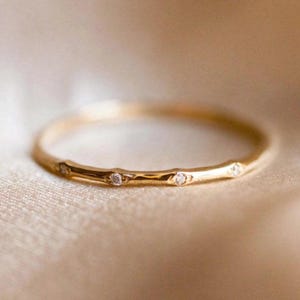 Könnte beinhalten: Ein zarter Goldring mit kleinen, funkelnden Diamanten, die gleichmäßig über das Band verteilt sind. Der Ring hat eine glatte, polierte Oberfläche und ist vor einem weichen, neutralen Hintergrund platziert. Dieses elegante Schmuckstück ist perfekt für den Alltag.
