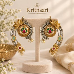 May include: A pair of gold-toned earrings featuring a central circular medallion with green and pink stones, surrounded by a crescent-shaped frame with lotus flower motifs, small white beads, and light green accents. The text Kritnaari is visible.
