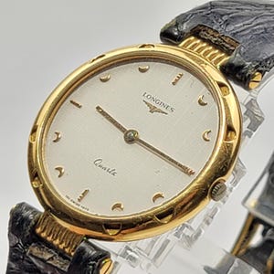 Puede incluir: Un reloj de pulsera de cuarzo Longines en tono dorado con caja redonda, bisel dorado texturizado, esfera blanca con patrón de cuadrícula sutil, manecillas doradas e índices en forma de media luna, con correa de cuero negro texturizado.