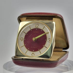 May include: A square gold-tone Seiko travel clock with a maroon centre dial and white outer ring. The clock features gold-tone hands, gold-tone numerals at the 3, 6, 9, and 12 positions, and the text Seiko and 2 Jewels printed on the face.