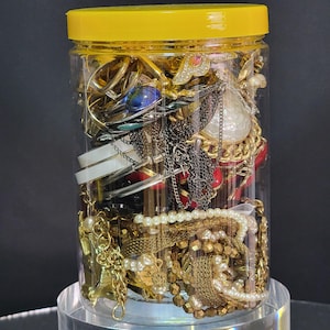 May include: A clear plastic jar with a bright yellow lid filled with a variety of assorted jewellery pieces. The collection includes gold-tone chains, white pearl-style beaded strands, metallic bangles, and various decorative charms and pendants.