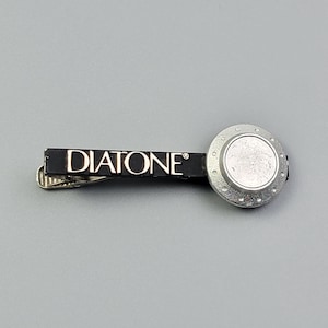 May include: A black tie clip featuring the word DIATONE in white lettering on the horizontal bar. The right side of the clip has a circular silver-toned accent with a textured border and a smooth, raised center disc, attached to a metal clasp.