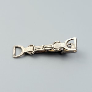May include: A silver-toned metal tie slide featuring a horizontal equestrian horse bit design. The accessory has a polished finish with two D-shaped ends connected by a central bar and two circular rings, secured with a spring-loaded clip mechanism.
