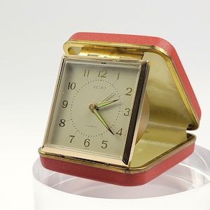 May include: A square Seiko travel alarm clock with a gold-tone metal frame and a cream-coloured face featuring black Arabic numerals. The clock is housed in a hinged, coral-red textured case with a gold-tone interior trim and luminous hands.
