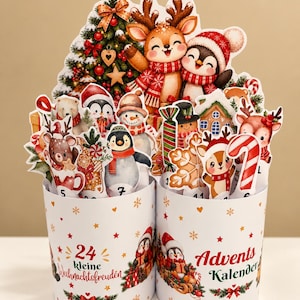 Könnte beinhalten: Ein Adventskalender in Form von zwei Zylindern mit einer festlichen Darstellung von Zeichentricktieren wie Rentieren, Pinguinen und einem Schneemann. Die weißen Behälter sind mit roten und goldenen Sternen, Schneeflocken und dem Text 24 kleine Weihnachtsfreuden Advents Kalender verziert.