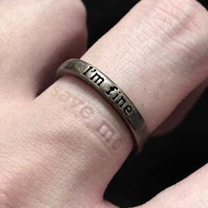 Peut inclure: Une bague de couleur bronze avec l'inscription "I'm fine" gravée à l'extérieur. La bague est portée au doigt, avec les mots "save me" faiblement visibles sur la peau en dessous. La bague a un design simple, de type anneau.