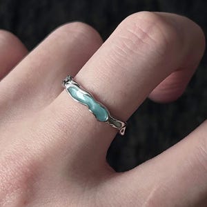 Puede incluir: Un anillo plateado con un diseño ondulado, con una incrustación de esmalte azul claro. El anillo se lleva en un dedo, mostrando su forma orgánica única y sus colores contrastantes. El diseño del anillo recuerda al agua que fluye.