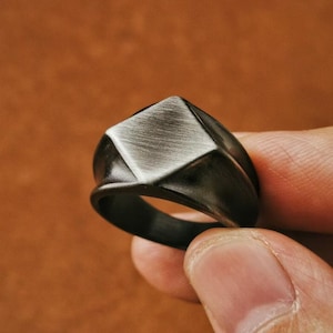 May include: A black ring with a geometric, diamond-shaped top. The top has a brushed metal finish, contrasting with the smooth, dark band. The ring is held in a hand, showcasing its design and texture.