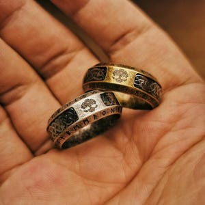May include: Two ornate rings with a tree of life design. One ring is gold-toned, the other is silver-toned. Both rings have intricate patterns and are likely made of metal. The rings are displayed on a hand.
