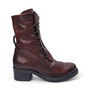 May include: A brown leather boot with a lace-up front and a side zipper. The boot has a short heel and a pull tab at the top. The leather has a distressed look, giving it a vintage feel. The boot is designed for everyday wear.
