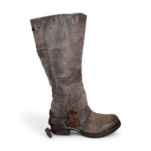 May include: A distressed brown leather knee-high boot with visible stitching and a metal spur. The boot features a worn, textured appearance, with a buckle and strap detail near the ankle. The overall design suggests a vintage or rustic aesthetic.