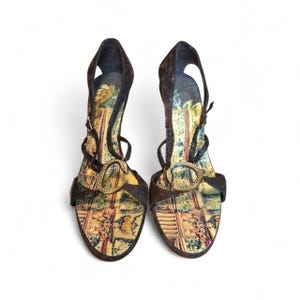 May include: A pair of high-heeled sandals featuring dark brown textured straps and a gold-toned letter C embellishment on the front. The footbed displays a colorful, detailed landscape print with trees and architectural elements in shades of green and tan.
