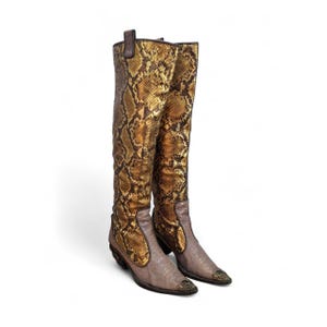 Könnte beinhalten: Ein Paar kniehohe Stiefel mit einem metallisch goldenen und braunen Schlangenledermuster am Schaft und einem Fußteil aus einfarbigem taupefarbenem Leder. Die Stiefel haben eine spitze Zehenpartie, einen Blockabsatz und eine Metallverzierung.