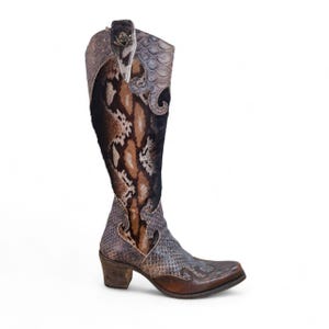 May include: A knee-high western boot featuring a mix of brown and grey snakeskin-textured leather, dark brown fur panels, and a stacked wooden heel. The boot includes decorative stitching, a pointed toe, and a metallic emblem on the upper shaft.